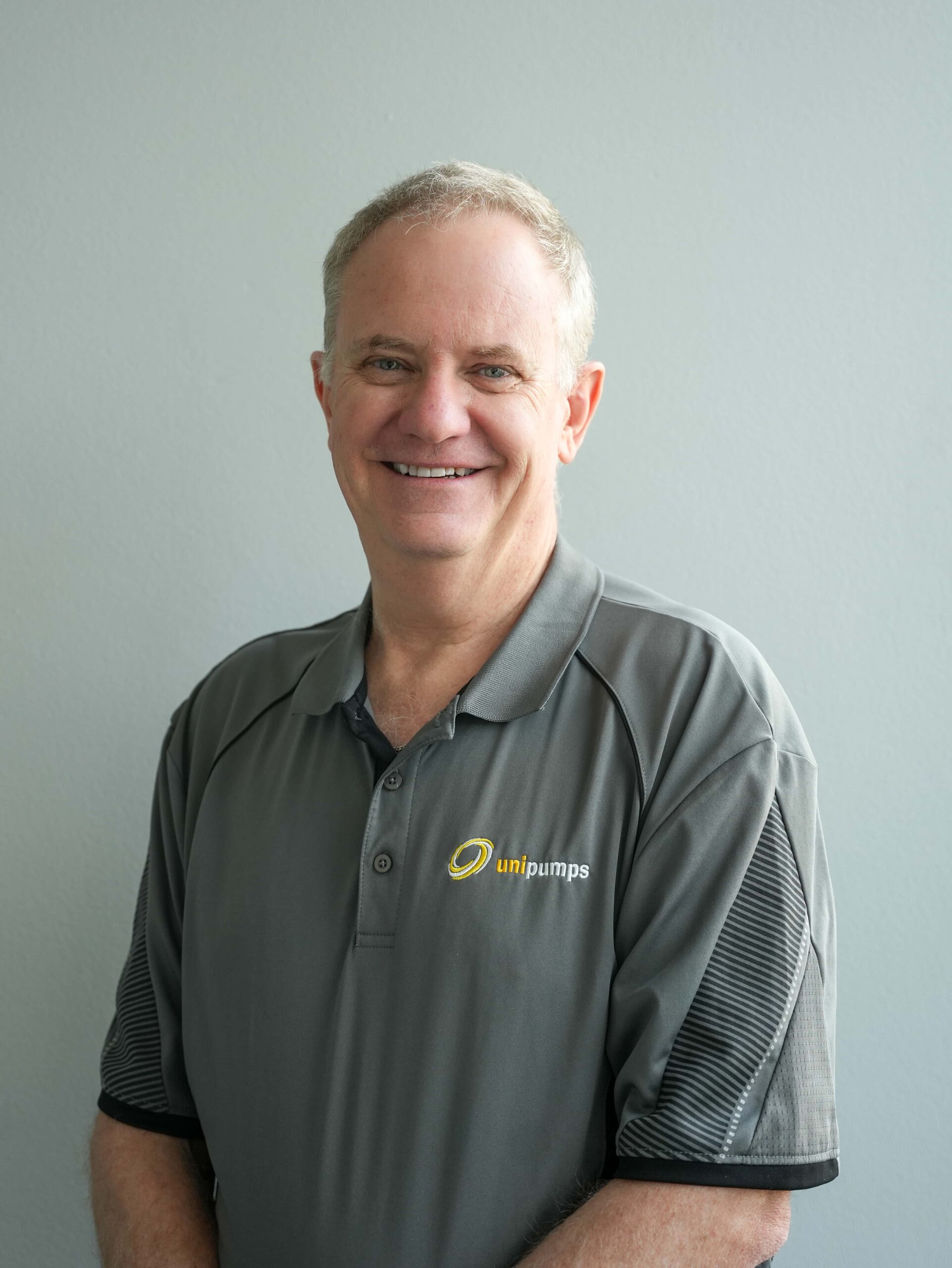 Andrew Rickard, smiling with short gray hair, wears a gray unipumps polo shirt by a light gray wall.